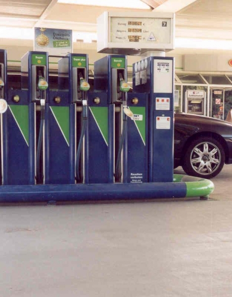 Mehrere grün-blaue Zapfsäulen an einer Tankstelle mit Auto im Hintergrund.